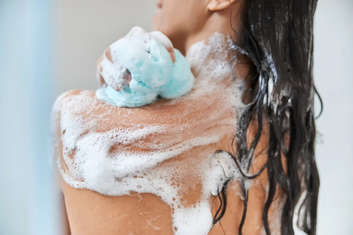 Woman using blue shower loofah with soap suds on back and wet hair in bathroom