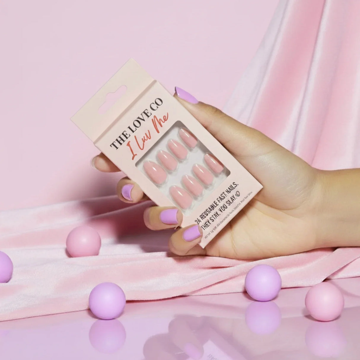 Hand holding The Love Co I Luv Me pink press on nails against pink satin and pastel balls