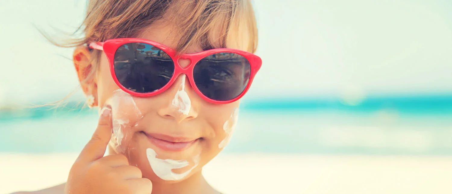 Child at beach wearing red sunglasses and applying sunscreen on face