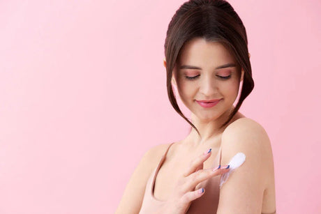 Woman applying body lotion on shoulder against pink background