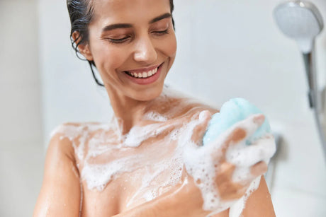 Smiling woman showering with blue loofah and soap suds in modern bathroom