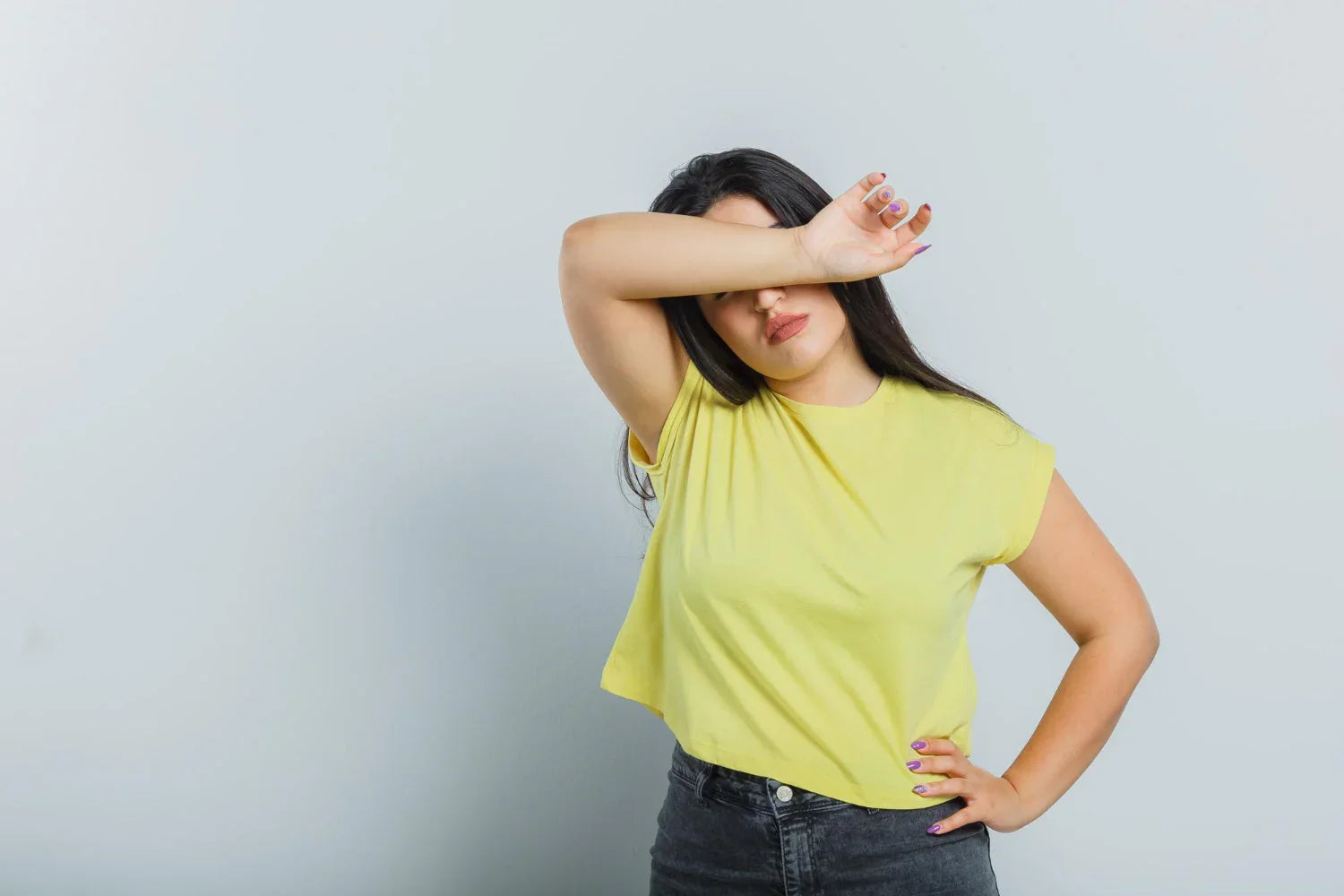 Woman in yellow t-shirt covering eyes with arm, standing against plain light background
