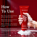 Hand holding The Love Co Fall In Love shower gel tube with soapy foam on a red background