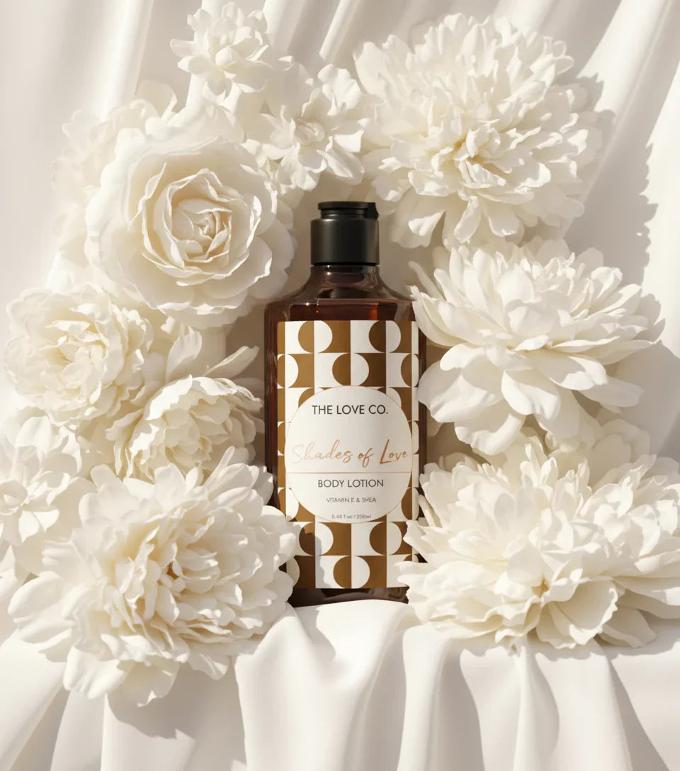 The Love Co. Shades of Love body lotion bottle with gold label, surrounded by white flowers on cream fabric