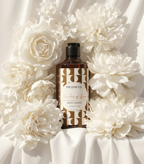 The Love Co. Shades of Love body lotion bottle with gold label, surrounded by white flowers on cream fabric