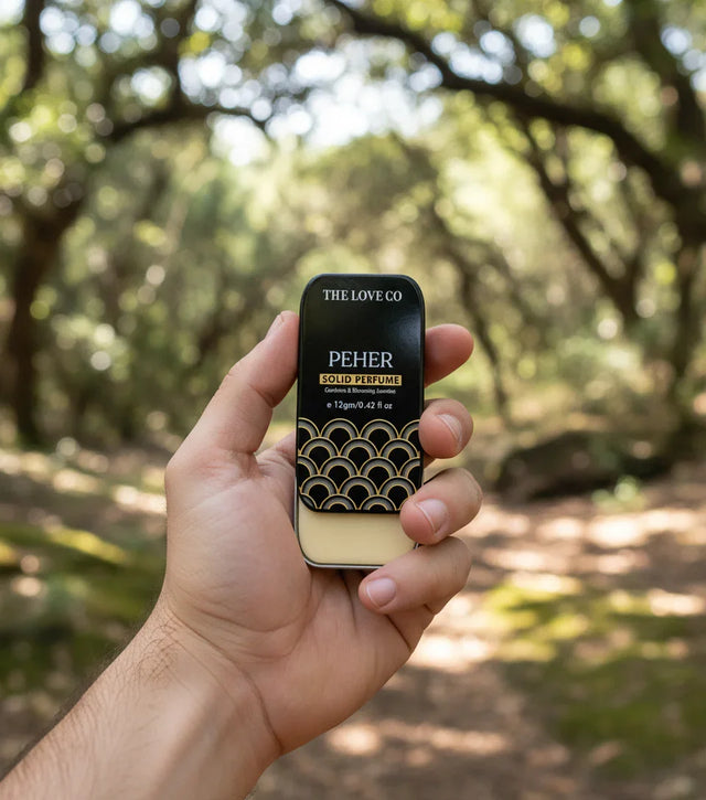 Hand holding The Love Co Peher solid perfume tin outdoors in a forest setting