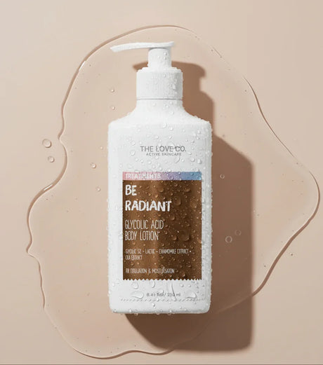 The Love Co. Be Radiant Glycolic Acid Body Lotion bottle with water droplets on a beige background.