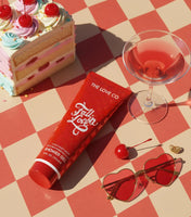 Red product packaging with 'The Love Co' branding on a checkered tablecloth with cake, glass, and sunglasses.