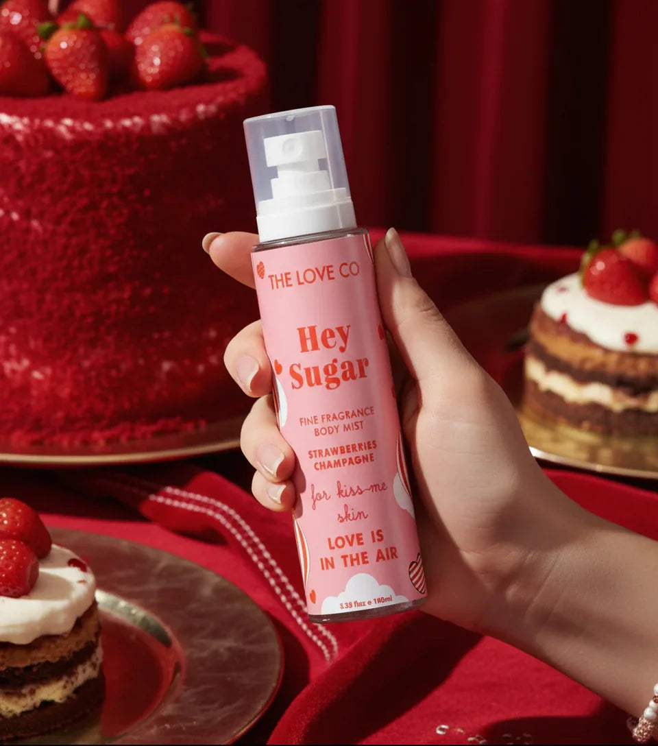 Hand holding The Love Co Hey Sugar body mist with strawberries and cake on a red tablecloth