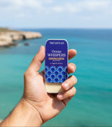Hand holding The Love Co Ocean Whispers solid perfume tin by the ocean
