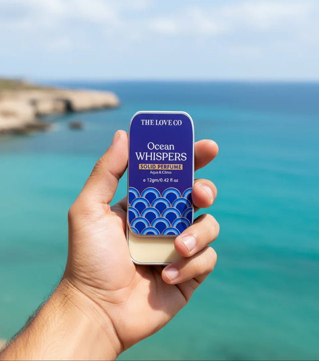 Hand holding The Love Co Ocean Whispers solid perfume tin by the ocean